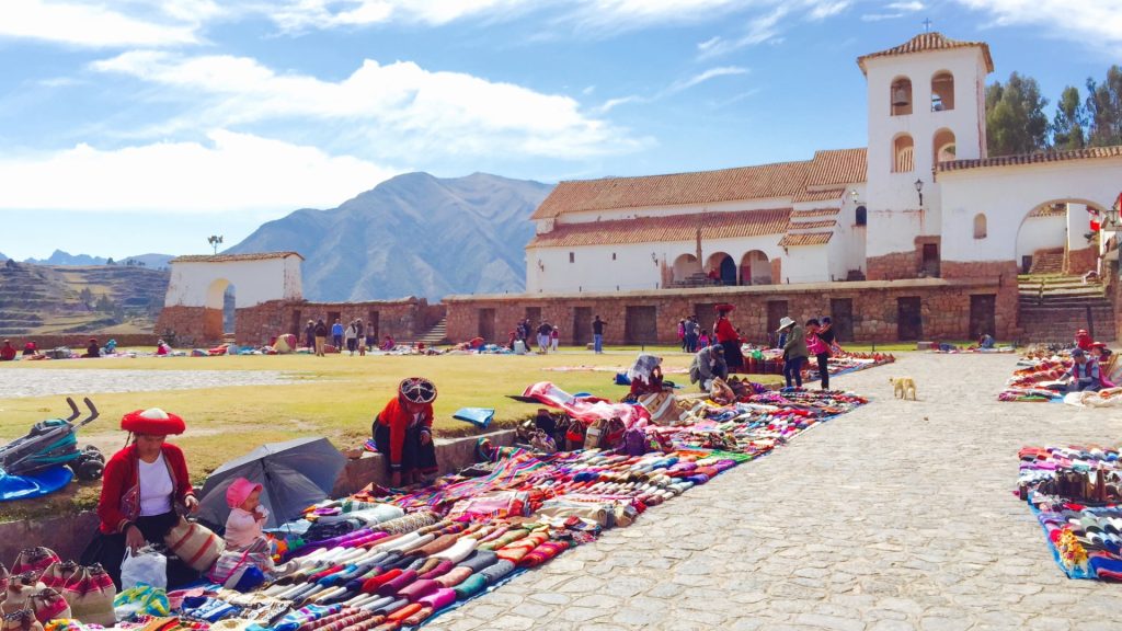 Andean vendors in traditional clothing selling handmade textiles in front of a colonial church during a sacred valley tour stop – Ali Peru Treks