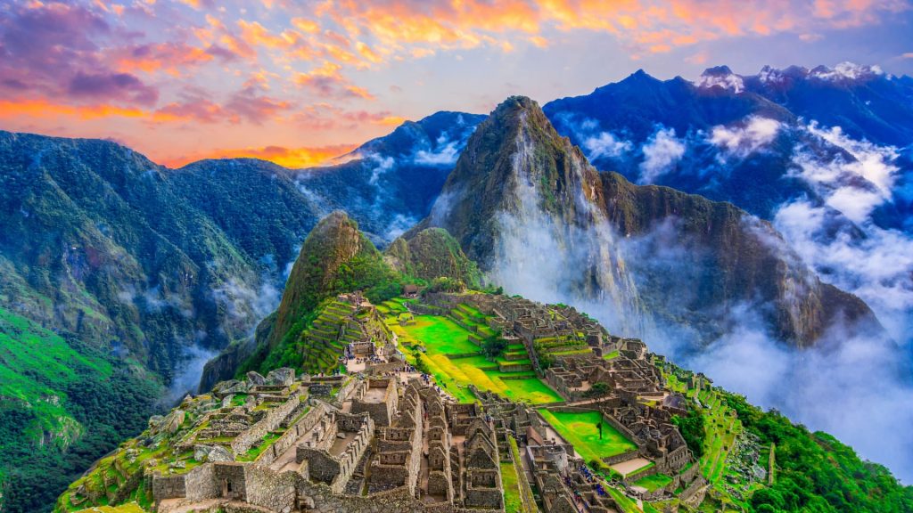 A breathtaking view of Machu Picchu at sunrise, with golden light illuminating the ancient Inca citadel, surrounded by lush mountains and misty clouds - Ali Peru Trek