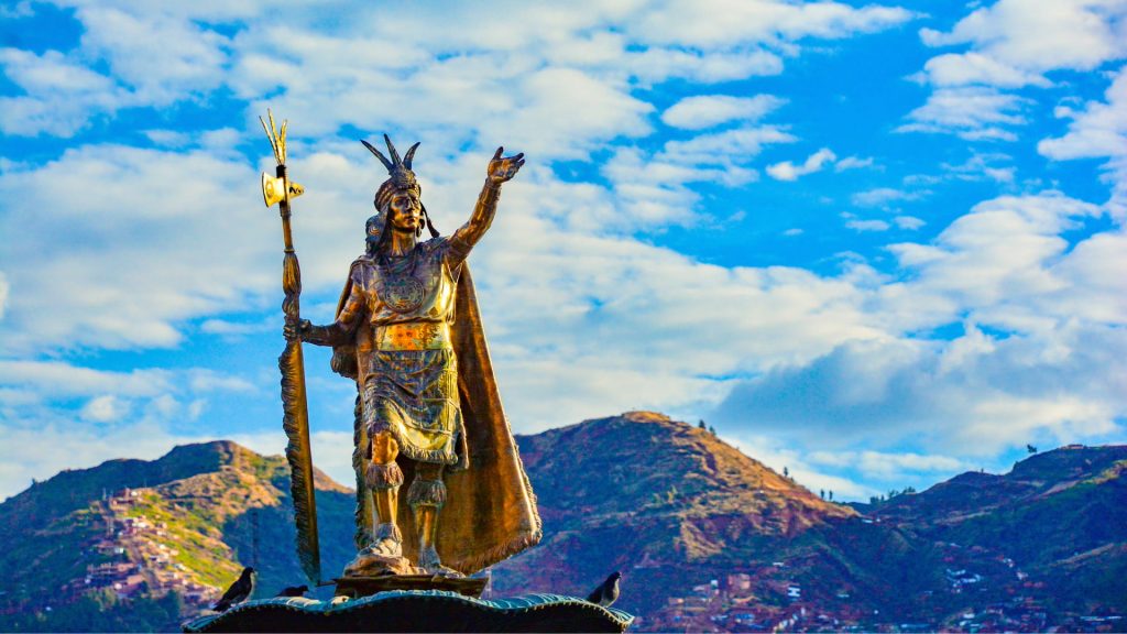Monument of Pachacútec in Cusco’s main square, depicting the great Inca emperor with his golden attire and staff, set against a backdrop of mountains and blue sky - Ali Peru Treks.