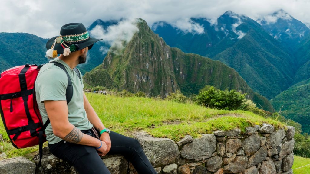 A traveler with a red backpack and Andean hat enjoys a breathtaking view of Machu Picchu, surrounded by lush mountains - Ali Peru Trek