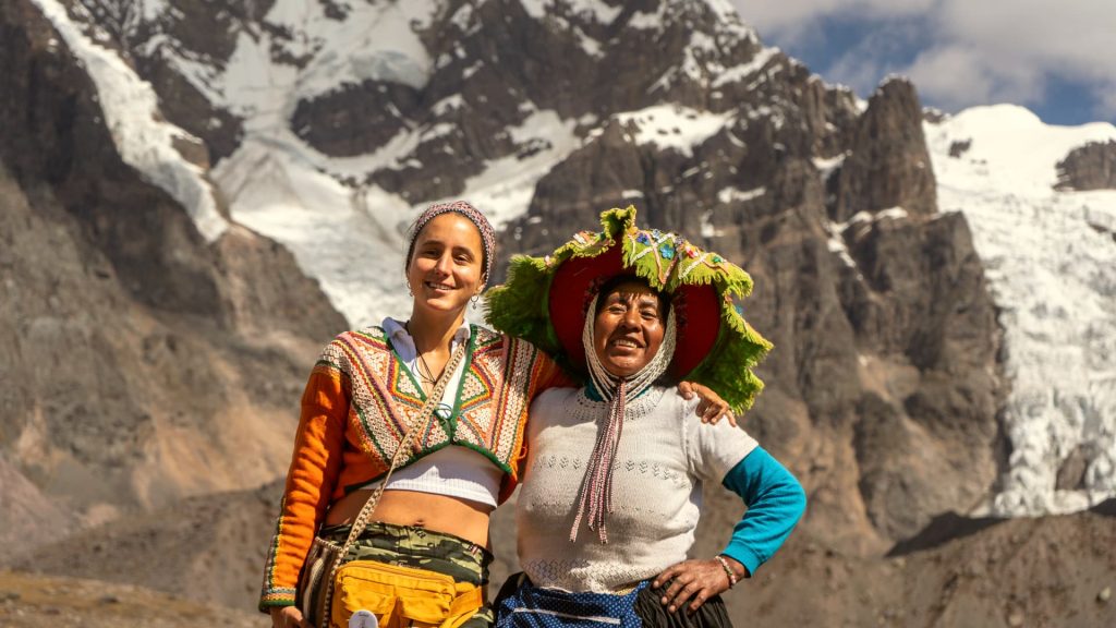 A traveler and an Andean woman in traditional clothing share a joyful moment in the Peruvian mountains, with stunning snow-capped peaks in the background. - Ali Peru Trek.