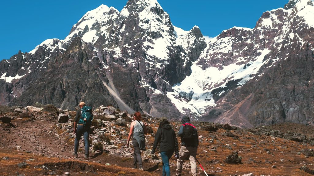 A group of trekkers hikes through the rugged Andean landscape, surrounded by towering snow-capped mountains under a bright blue sky. - Ali Peru Trek.