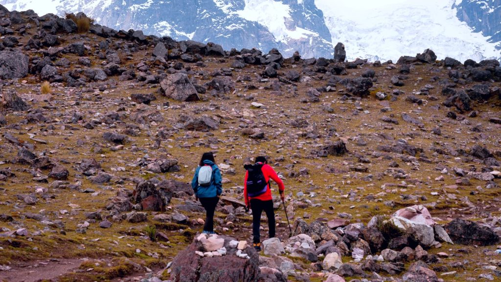 Two hikers traverse a rugged Andean trail in Peru, surrounded by rocky terrain with snow-capped peaks in the background. - Ali Peru Trek.
