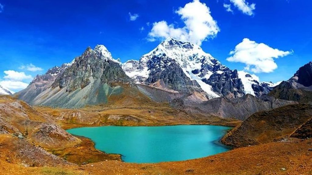 A stunning turquoise lake nestled in the Peruvian Andes, surrounded by towering snow-capped peaks and rugged mountain landscapes under a bright blue sky. - Ali Peru Trek.