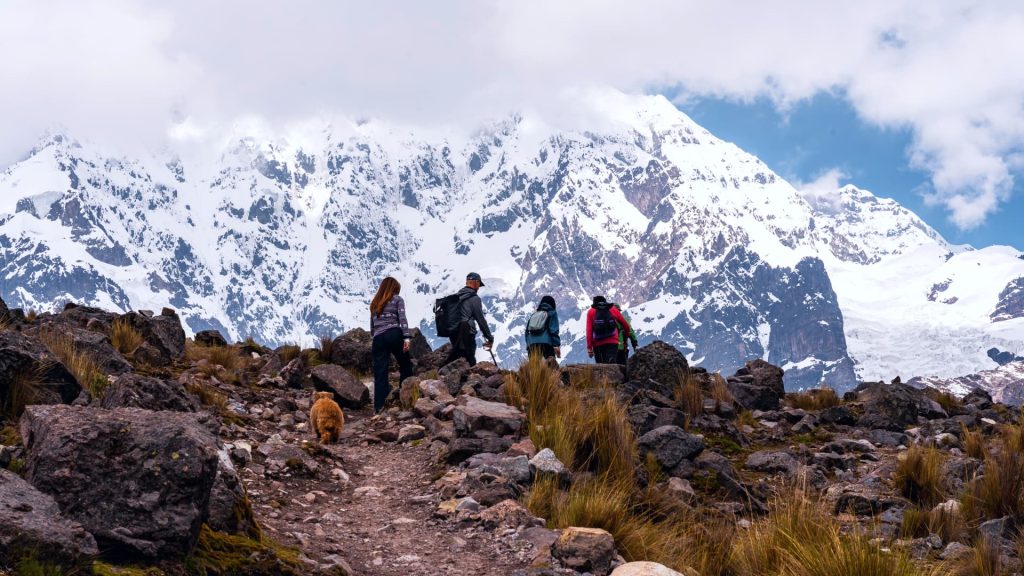 A group of hikers, accompanied by a dog, treks through the rugged Andean landscape towards majestic snow-capped peaks in Peru. - Ali Peru Trek.