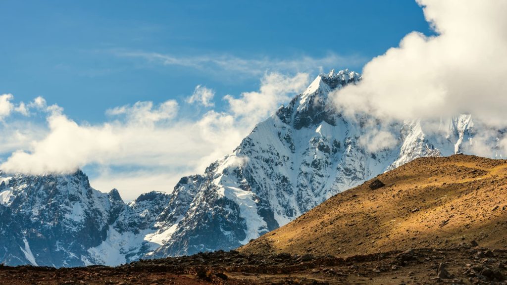 Majestic snow-capped peaks of the Peruvian Andes rise above rugged terrain, with clouds wrapping around the summits under a bright blue sky. - Ali Peru Trek.