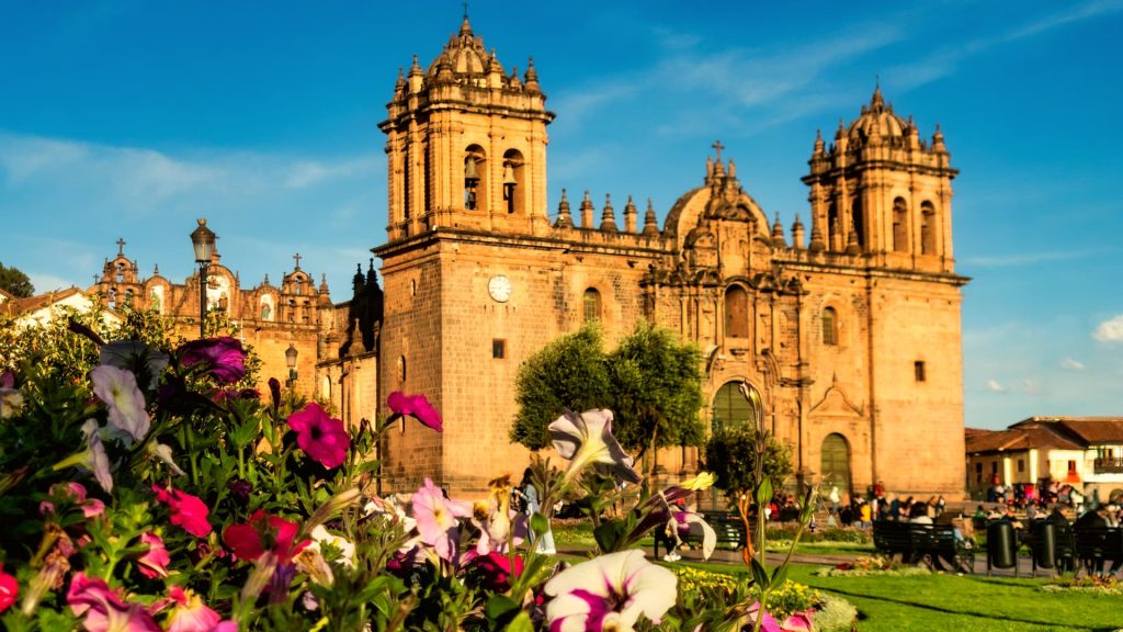 The Cusco Cathedral at sunset, bathed in golden light, with vibrant flowers in the foreground, capturing the charm of the Plaza de Armas in Peru. - Ali Peru Trek.