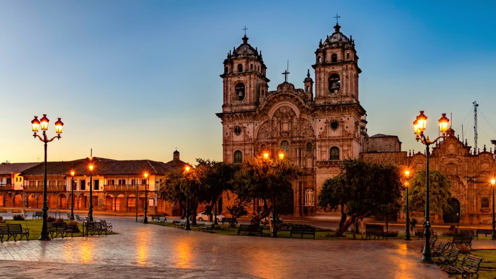 The San Francisco Church in Cusco, Peru, illuminated by warm streetlights at sunset, showcasing its colonial architecture in a peaceful plaza setting. - Ali Peru Trek.