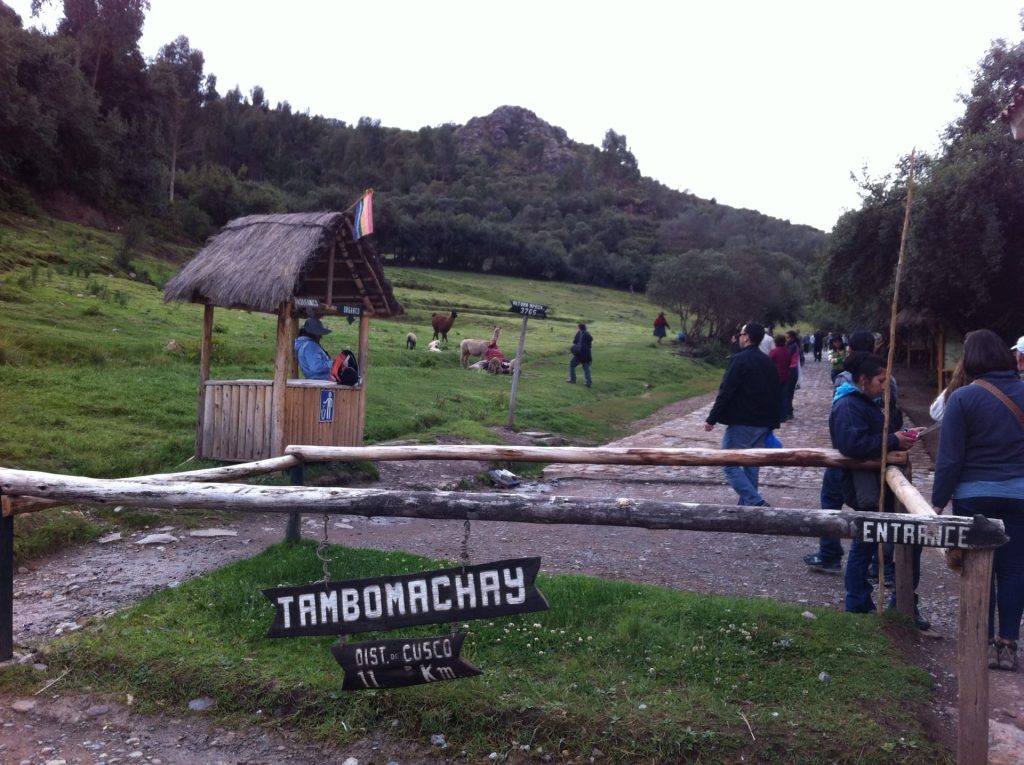 Entrance to the Tambomachay archaeological site in Cusco, featuring a welcome sign, visitors, and Andean landscape. - Ali Peru Treks