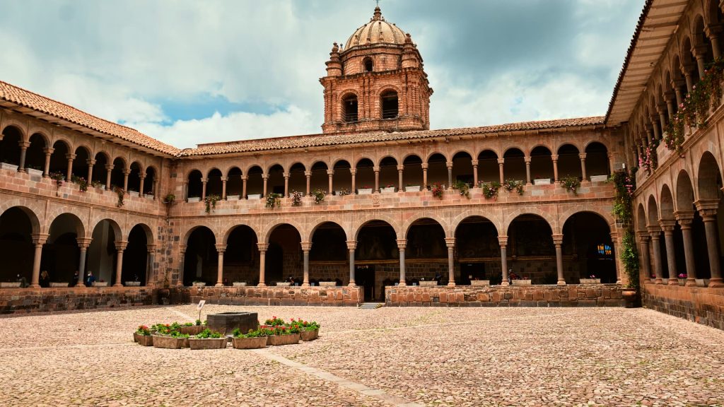 The colonial courtyard of Qorikancha in Cusco, Peru, featuring elegant arches, a central fountain, and the stunning bell tower of the Santo Domingo Church. - Ali Peru Trek.