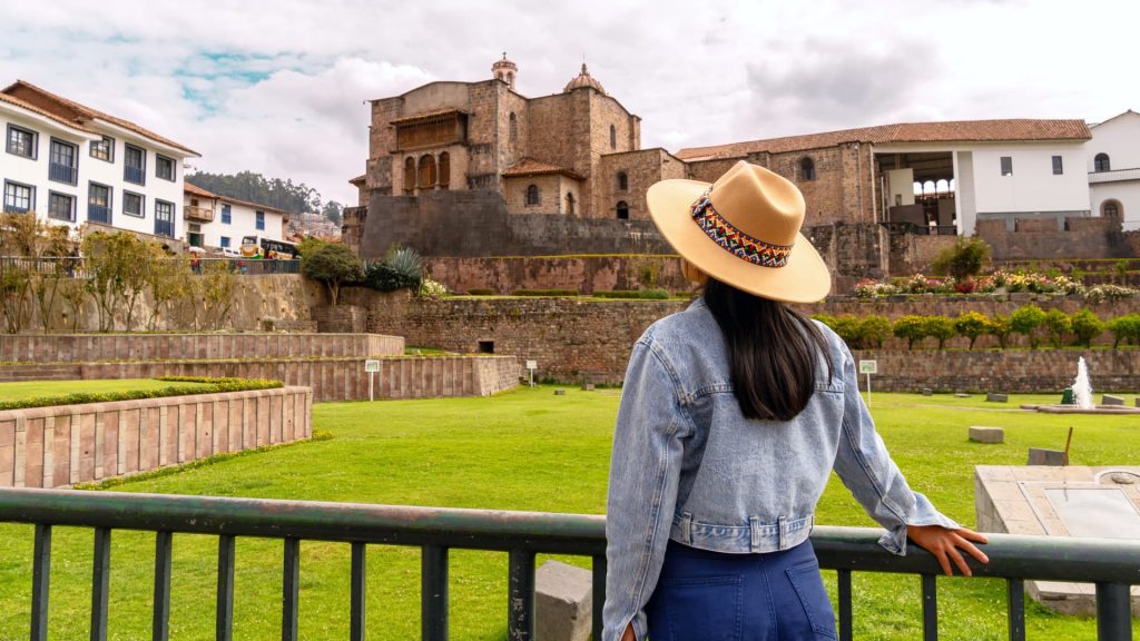 A female traveler in a hat and denim jacket admires the historic Qorikancha temple in Cusco, Peru, surrounded by lush gardens and Inca stone walls. - Ali Peru Trek.