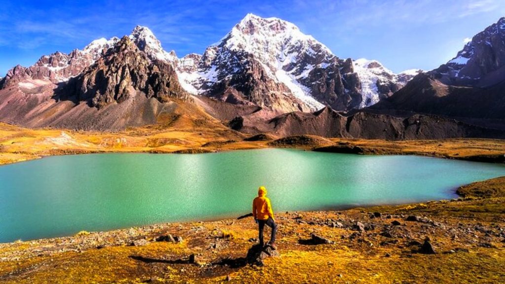 A traveler in a yellow jacket admires a stunning turquoise lake in the Peruvian Andes, surrounded by snow-capped mountains and golden highland terrain. - Ali Peru Trek.