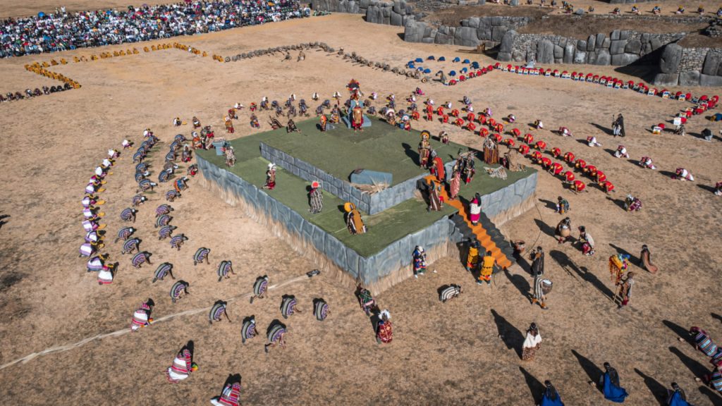 Overhead view of the Inti Raymi festival re-enactment at Sacsayhuamán, showing performers in colorful traditional costumes arranged in formations around a central platform, with large crowds surrounding the area - Ali Peru Treks.