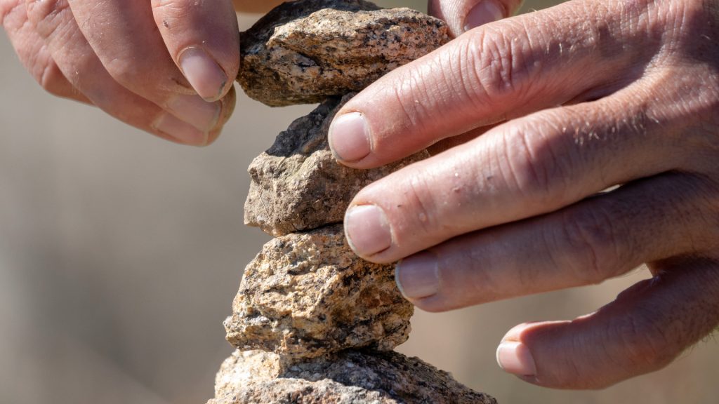 Close-up of hands carefully placing a rock on top of other stacked stones, in the process of building a small stone cairn, or Apacheta, against a blurred natural background - Ali Peru Treks.