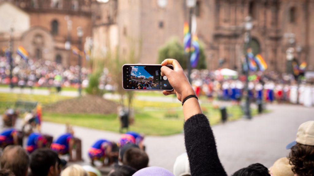 A person's hand holding a smartphone, filming a vibrant festival or procession in the main square of Cusco, with traditional buildings and crowds in the background, possibly during Inti Raymi celebrations - Ali Peru Treks.