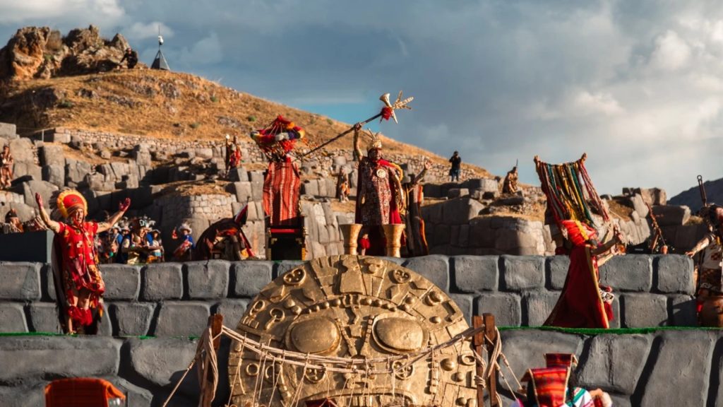 Performers in elaborate traditional Inca costumes re-enact a central ritual of the Inti Raymi festival on a stone stage with a large golden sun disk, set against the ancient walls of Sacsayhuamán - Ali Peru Treks.