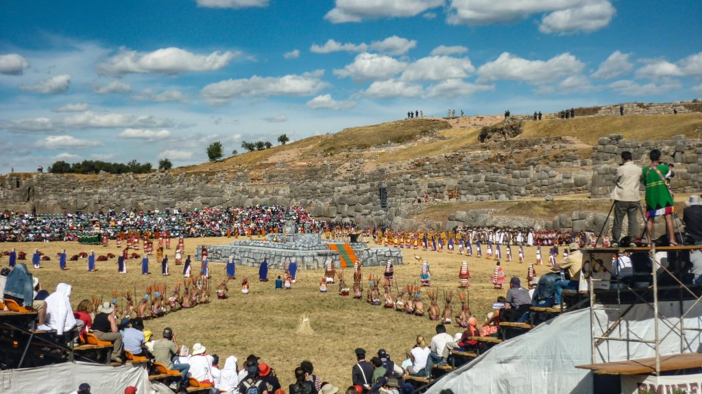 Panoramic view of the grand Inti Raymi festival re-enactment at the Sacsayhuamán archaeological complex, showing performers in traditional costumes and a large audience gathered under a cloudy sky - Ali Peru Treks.