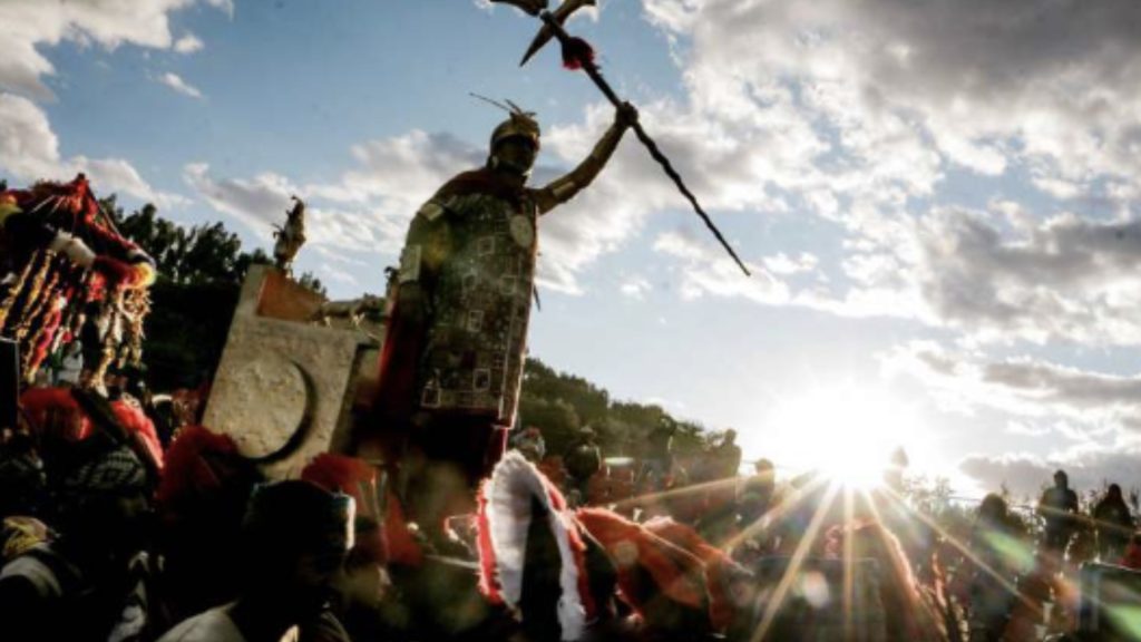 An actor portraying the Inca Sapa in ceremonial attire raising a staff during the Inti Raymi festival, with the sun setting brightly in the background - Ali Peru Treks.