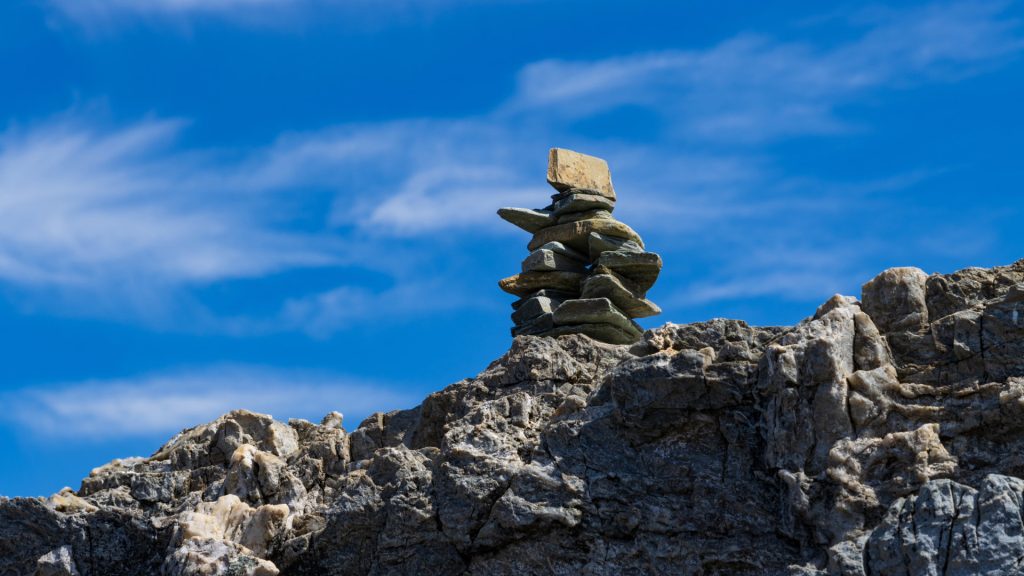 A well-balanced stack of rocks, known as the Apacheta, perched on a rugged mountain peak under a bright blue sky, symbolizing a trail marker or offering - Ali Peru Treks.
