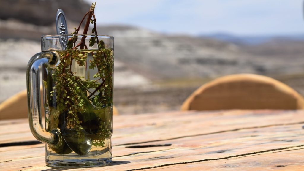 Glass mug with coca and muña leaves steeping in hot water, served outdoors on a rustic wooden table with mountain views – Ali Peru Treks