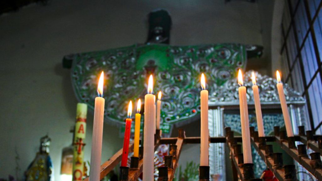 Rows of burning candles in front of a religious figure dressed in green, reflecting the ceremonial ambiance of Cruz Velacuy – Ali Peru Treks