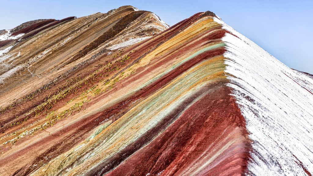 A wide panoramic landscape of the uniquely colored geological strata of Rainbow Mountain, showing its vibrant yellow, red, and brown hues under a dramatic sky. - Ali Peru Treks