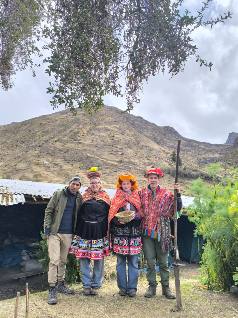 A group of people posing together in front of a mountainous landscape in the Andes, dressed in a mix of modern hiking gear and colorful traditional Peruvian textiles