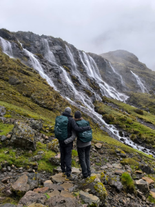 The scene shows two people admiring the 7 Quishuarani Waterfalls in the district of Lares