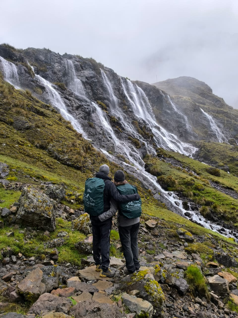 The scene shows two people admiring the 7 Quishuarani Waterfalls in the district of Lares